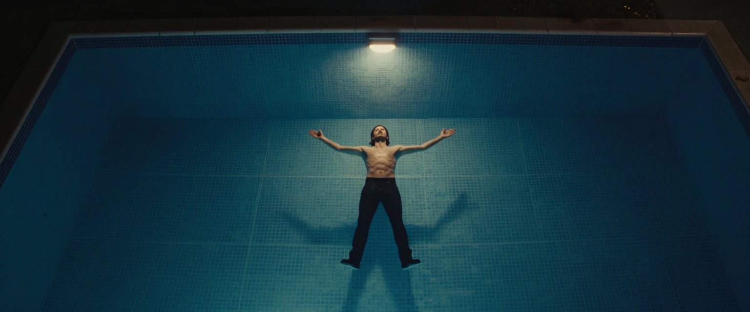 Deep End: a singer lying on the bottom of an empty tiled swimming pool at night, arms spread, lit from above by a single cold light